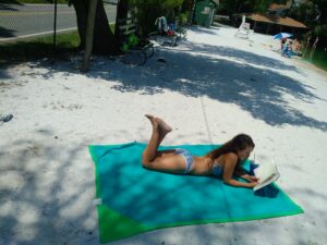 A woman reading on a green towel at a sandy beach.
